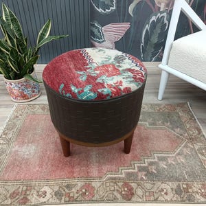May include: A round, brown leather ottoman with a colorful patterned top. The ottoman is sitting on a red and beige rug.