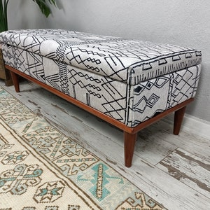May include: A white and black patterned storage bench with wooden legs. The bench features a geometric design and is ideal for adding a touch of style to any room.