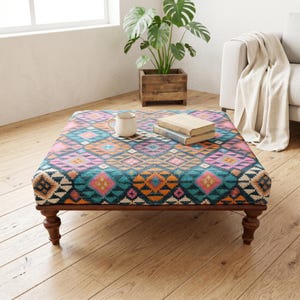 May include: Square ottoman with a colorful geometric pattern in teal, pink, orange, and white. The wooden frame has turned legs. A white mug and two books sit on top of the ottoman. A potted plant and a sofa are in the background.