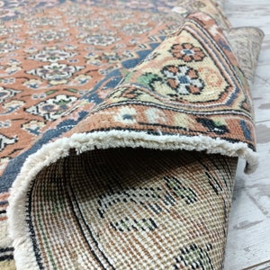 May include: A close-up of a hand-woven rug with a floral pattern in shades of pink, blue, and cream. The rug has a fringe border and is folded in half, revealing the back side.