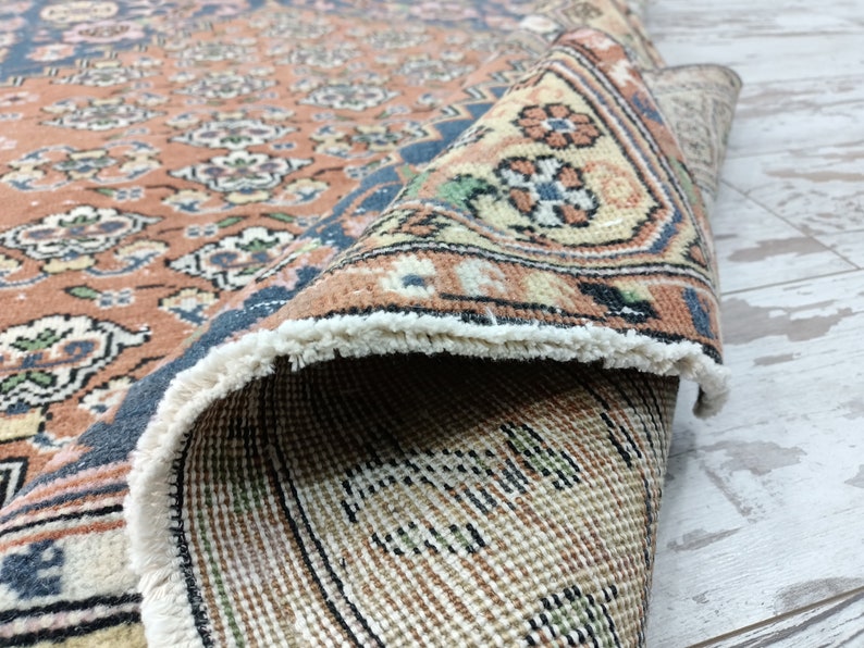 May include: A close-up of a hand-woven rug with a floral pattern in shades of pink, blue, and cream. The rug has a fringe border and is folded in half, revealing the back side.