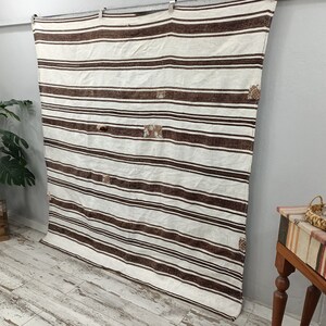 Puede incluir: Un textil rectangular grande de color crema con rayas horizontales marrones. El textil tiene una textura tejida y varios parches de costura decorativa. Est&aacute; colgado contra una pared gris.
