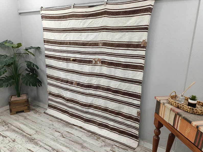 Puede incluir: Un textil rectangular grande de color crema con rayas horizontales marrones. El textil tiene una textura tejida y varios parches de costura decorativa. Est&aacute; colgado contra una pared gris.