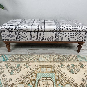 May include: A black and white patterned storage ottoman with wooden legs. The ottoman is sitting on a cream-coloured rug with a blue and brown geometric pattern.