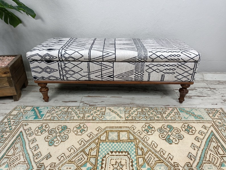 May include: A black and white patterned storage ottoman with wooden legs. The ottoman is sitting on a cream-coloured rug with a blue and brown geometric pattern.