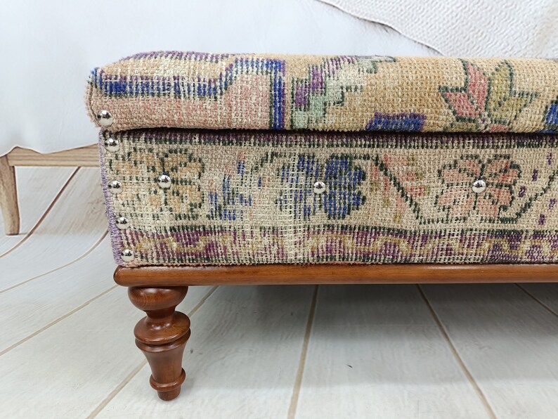 May include: A rectangular ottoman with a floral patterned fabric top and wooden legs. The fabric is a beige, pink, and blue floral pattern with a dark brown border. The legs are a dark brown wood.