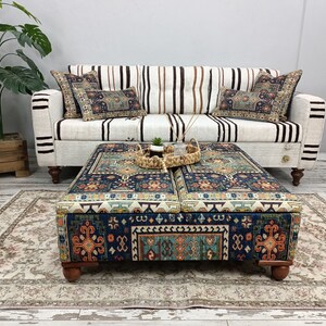 May include: A patterned ottoman with a dark blue, orange, and green design. The ottoman has four wooden legs and is sitting on a cream-colored rug in front of a white and brown striped sofa.