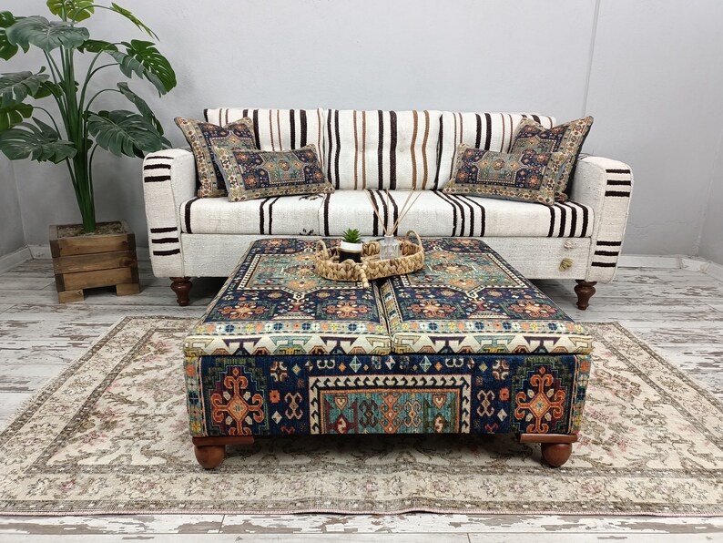 May include: A patterned ottoman with a dark blue, orange, and green design. The ottoman has four wooden legs and is sitting on a cream-colored rug in front of a white and brown striped sofa.