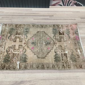 May include: A small, rectangular rug with a faded, vintage design. The rug has a light beige background with a central medallion in shades of green and brown. The edges of the rug are frayed and worn, giving it a rustic look.