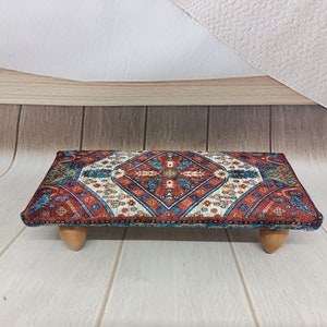 May include: A small, rectangular footstool with a colorful, patterned fabric top and wooden legs. The fabric features a geometric design in shades of red, blue, and white.