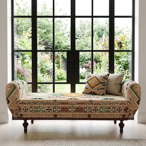 May include: A patterned bench in beige, green, and orange, with two cushions. The bench has curved arms and dark wooden legs. The fabric features geometric patterns. The background shows a large window with a view of a garden. The bench is approximately 150 cm long.