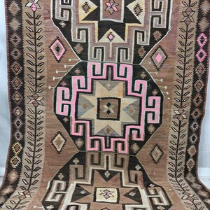 May include: A vintage Turkish kilim rug with a geometric pattern in shades of brown, pink, and white. The rug has a central medallion with a starburst design and is surrounded by a border of repeating motifs.
