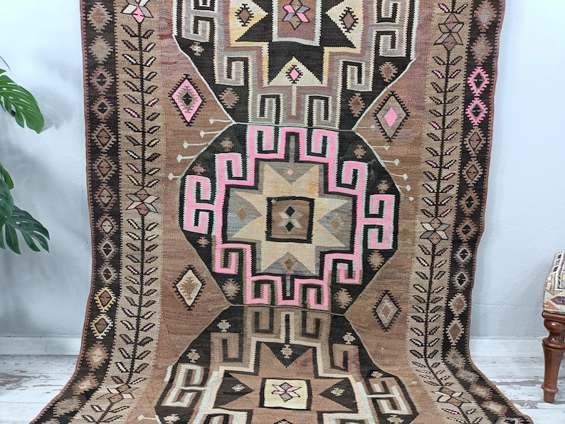 May include: A vintage Turkish kilim rug with a geometric pattern in shades of brown, pink, and white. The rug has a central medallion with a starburst design and is surrounded by a border of repeating motifs.