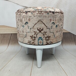 May include: A round ottoman with a beige fabric cover featuring a brown, blue, and green floral pattern. The ottoman has four white wooden legs.