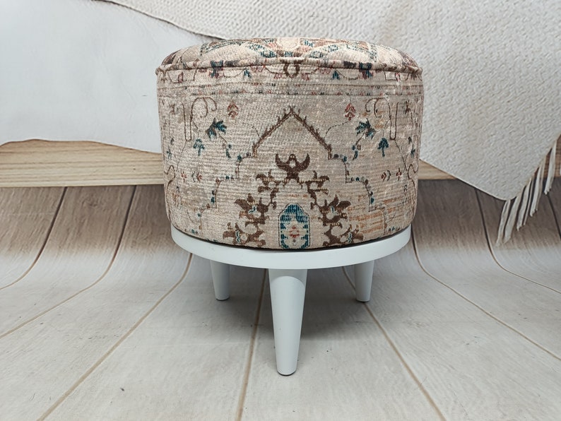 May include: A round ottoman with a beige fabric cover featuring a brown, blue, and green floral pattern. The ottoman has four white wooden legs.