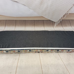 Pu&ograve; includere: Un cuscino rettangolare grigio e bianco con un bordo a motivi. Il cuscino &egrave; su un pavimento di legno.