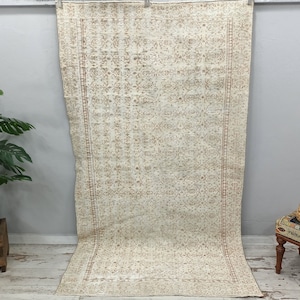 Puede incluir: Una alfombra vintage de color crema con un patr&oacute;n floral marr&oacute;n. La alfombra tiene un borde con flecos y est&aacute; colgada en una pared.