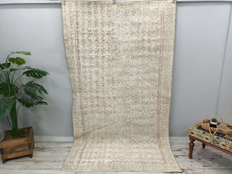 Puede incluir: Una alfombra vintage de color crema con un patr&oacute;n floral marr&oacute;n. La alfombra tiene un borde con flecos y est&aacute; colgada en una pared.