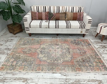 Puede incluir: Una alfombra vintage con un patrón floral descolorido en rosa y gris sobre un fondo blanco. La alfombra está colocada sobre un suelo de madera blanqueada en frente de un sofá blanco con cojines a rayas.