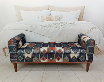 May include: A patterned storage bench with a wooden frame and legs. The bench features a dark blue fabric with geometric patterns in orange, white, and brown. The bench is placed in front of a bed with white bedding.