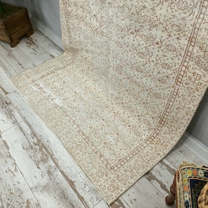 Puede incluir: Una alfombra de color crema con un delicado patr&oacute;n floral en color marr&oacute;n claro. La alfombra tiene un borde con un patr&oacute;n repetitivo. La alfombra est&aacute; colocada sobre un suelo de madera blanqueada.