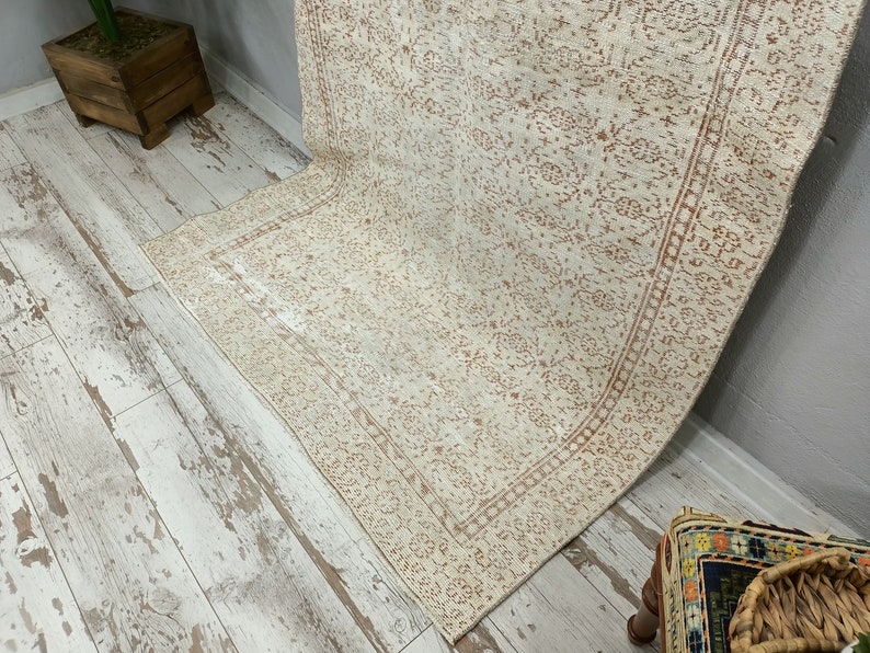 Puede incluir: Una alfombra de color crema con un delicado patr&oacute;n floral en color marr&oacute;n claro. La alfombra tiene un borde con un patr&oacute;n repetitivo. La alfombra est&aacute; colocada sobre un suelo de madera blanqueada.