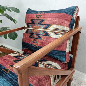 K&ouml;nnte beinhalten: Holzsessel mit Kissen und Sitzfl&auml;che mit einem von amerikanischen Ureinwohnern inspirierten Muster in Rot-, Blau- und Cremet&ouml;nen. Der Stuhl hat ein dunkles Holzgestell und steht vor einer Pflanze.