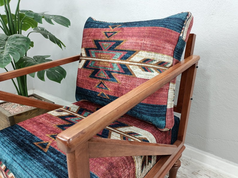 K&ouml;nnte beinhalten: Holzsessel mit Kissen und Sitzfl&auml;che mit einem von amerikanischen Ureinwohnern inspirierten Muster in Rot-, Blau- und Cremet&ouml;nen. Der Stuhl hat ein dunkles Holzgestell und steht vor einer Pflanze.