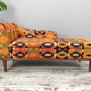 Puede incluir: Una chaise longue con estampado colorido y patas de madera. La chaise longue tiene un patrón geométrico en tonos naranja, amarillo, negro y verde.