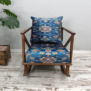 A blue patterned armchair with a wooden rocking chair next to it, placed on a white wooden floor. There is a potted plant to the left of the armchair.