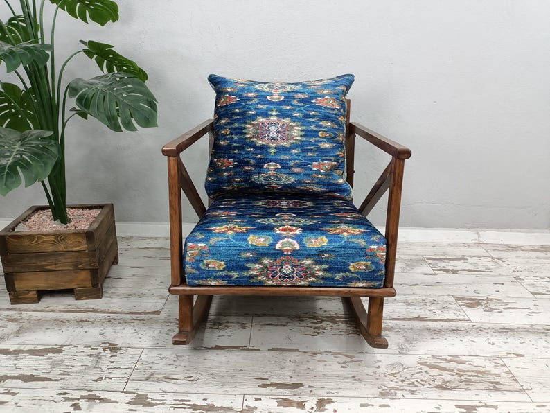 A blue patterned armchair with a wooden rocking chair next to it, placed on a white wooden floor. There is a potted plant to the left of the armchair.