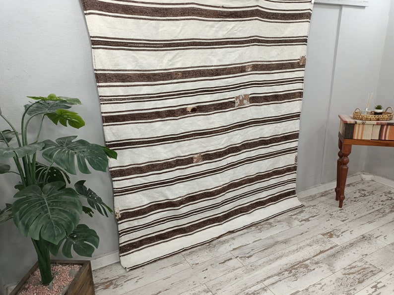 Puede incluir: Un textil a rayas blancas y marrones, posiblemente una alfombra o manta, cuelga verticalmente. El dise&ntilde;o presenta rayas horizontales marrones oscuras espaciadas uniformemente sobre un fondo blanco. Se ven parches de costura decorativa. El textil se exhibe en interiores.