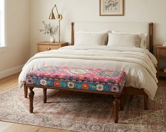 May include: A bedroom scene featuring a bed with a cream-colored headboard and bedding. A decorative bench with a pink, blue, and floral patterned cushion sits at the foot of the bed. The bench has dark brown wooden legs. A patterned rug and wooden nightstands complete the scene.