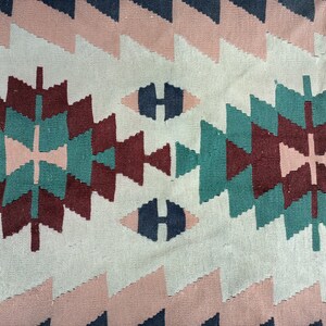 K&ouml;nnte beinhalten: Nahaufnahme eines gewebten Textils mit geometrischem Muster. Das Design zeigt sich wiederholende Formen in Rosa-, T&uuml;rkis-, Burgunder- und Marineblaut&ouml;nen vor einem cremefarbenen Hintergrund. Das Textil hat ein strukturiertes Aussehen.