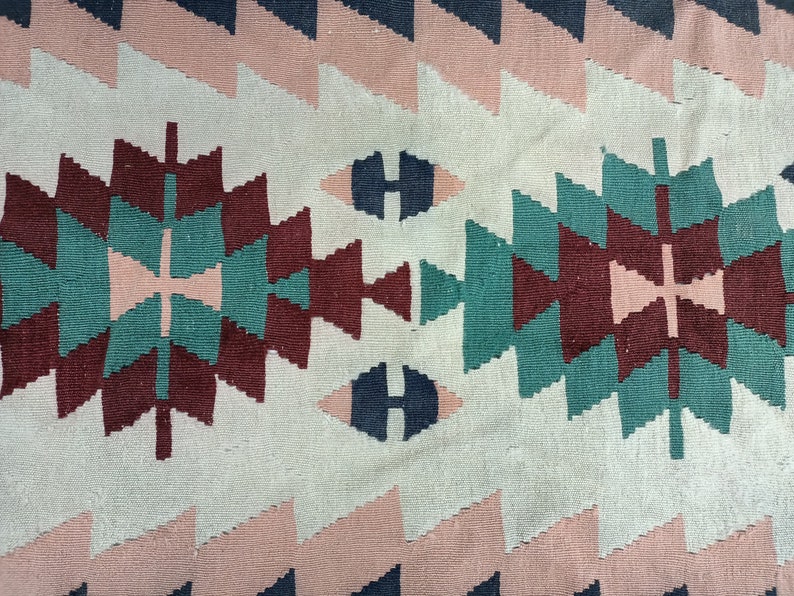 K&ouml;nnte beinhalten: Nahaufnahme eines gewebten Textils mit geometrischem Muster. Das Design zeigt sich wiederholende Formen in Rosa-, T&uuml;rkis-, Burgunder- und Marineblaut&ouml;nen vor einem cremefarbenen Hintergrund. Das Textil hat ein strukturiertes Aussehen.