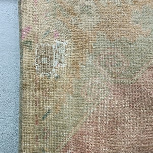May include: Close-up of a beige, pink, and green woven rug with a small hole in the centre. The rug has a textured surface and a frayed edge.