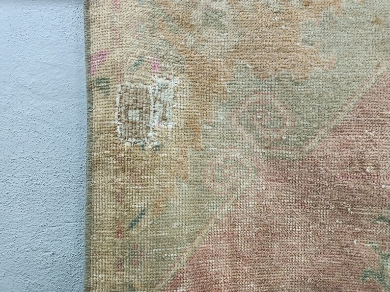 May include: Close-up of a beige, pink, and green woven rug with a small hole in the centre. The rug has a textured surface and a frayed edge.