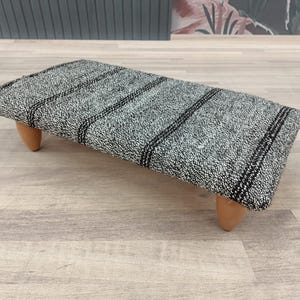 May include: A rectangular footstool with a gray and black striped fabric top. The footstool is supported by four short, rounded wooden legs. The fabric has a textured appearance, and the design is simple and modern.