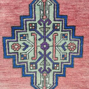 May include: A close-up of a red rug with a detailed blue, green, and purple geometric pattern in the center.
