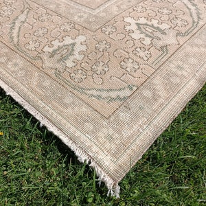 May include: Close-up of the corner of a beige and green patterned rug with a fringed edge. The rug is lying on a bed of green grass.