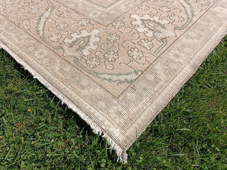 May include: Close-up of the corner of a beige and green patterned rug with a fringed edge. The rug is lying on a bed of green grass.
