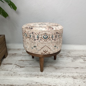 May include: A round ottoman with a beige fabric cover featuring a colourful floral pattern. The ottoman has four wooden legs.