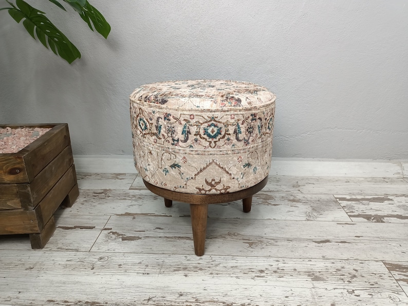 May include: A round ottoman with a beige fabric cover featuring a colourful floral pattern. The ottoman has four wooden legs.