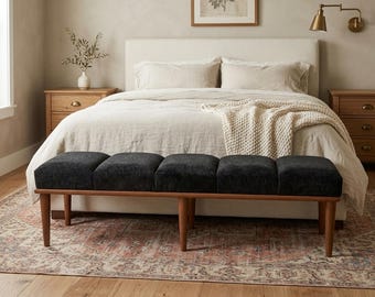 May include: A dark gray upholstered bench with a wooden frame and legs. The bench is placed at the foot of a bed with a cream-colored headboard and bedding. The room has a neutral color palette with a rug and wooden nightstands.