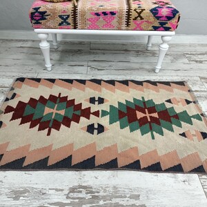 K&ouml;nnte beinhalten: Ein rechteckiger Teppich mit einem geometrischen Muster in Beige, Rosa, T&uuml;rkis und Burgunderrot. Der Teppich hat ein zentrales Rautenmuster und Zickzack-Bord&uuml;ren. Im Hintergrund befindet sich eine Bank mit wei&szlig;en Beinen und gemusterter Oberseite.