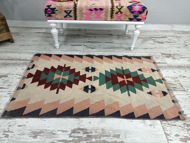K&ouml;nnte beinhalten: Ein rechteckiger Teppich mit einem geometrischen Muster in Beige, Rosa, T&uuml;rkis und Burgunderrot. Der Teppich hat ein zentrales Rautenmuster und Zickzack-Bord&uuml;ren. Im Hintergrund befindet sich eine Bank mit wei&szlig;en Beinen und gemusterter Oberseite.