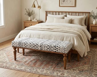 May include: A bedroom scene featuring a wooden bed frame, neutral-toned bedding, and a patterned bench at the foot of the bed. The bench has a black and white geometric design. A rug with a faded, vintage look sits beneath the bed and bench.