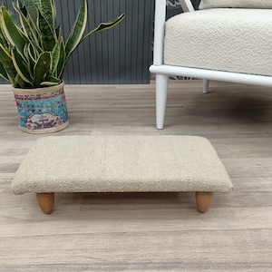 May include: A rectangular, beige footstool with a textured surface and four short, rounded wooden legs. The footstool is placed on a light-colored wooden floor. A potted plant and a white chair are in the background.