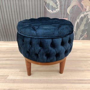 Puede incluir: Un otomano redondo de terciopelo azul oscuro con patas de madera. El otomano tiene una parte superior redonda y un diseño de capitoné con botones.