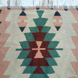 K&ouml;nnte beinhalten: Ein gewebter Teppich mit einem geometrischen Muster in Beige-, Rosa-, Gr&uuml;n- und Dunkelblaut&ouml;nen. Das Design zeigt sich wiederholende Dreiecks- und Diamantformen, die eine strukturierte Oberfl&auml;che erzeugen. Der Teppich hat einen Fransenrand.